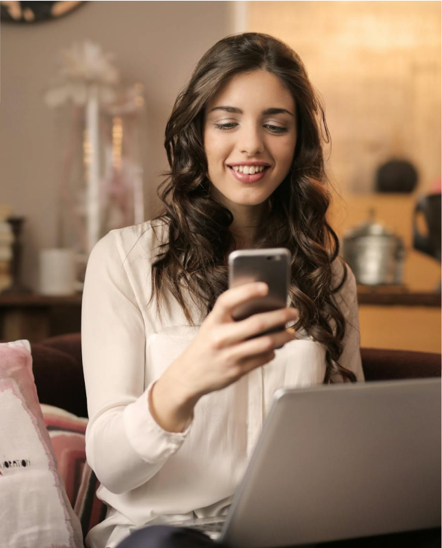 Smiling young woman looking at her phone while using a laptop, enjoying a productive or happy moment